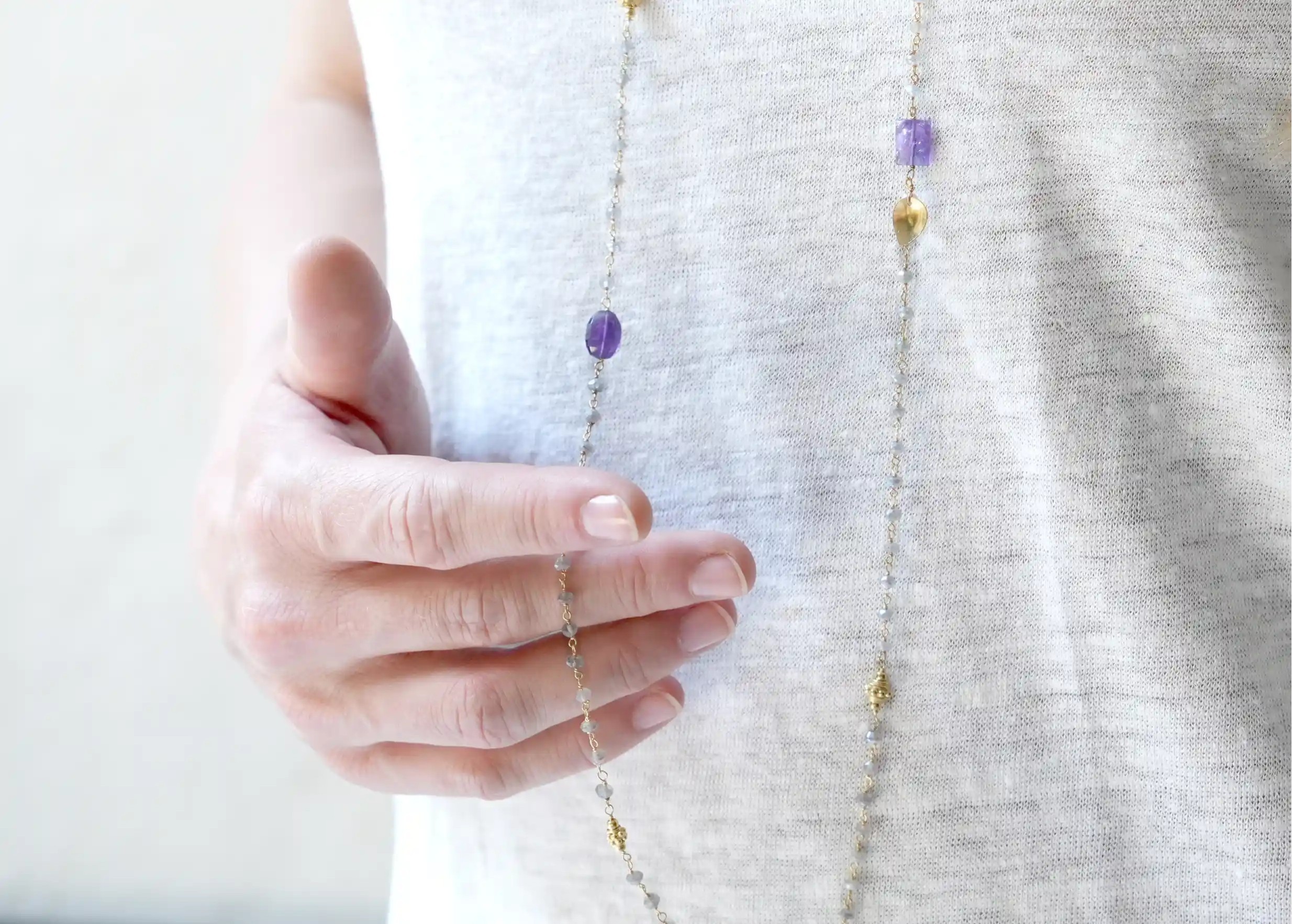 Femme tenant du bout de ses doigts un sautoir « Ani » Or & pierres d'améthyste et labradorite, collection Yamuna