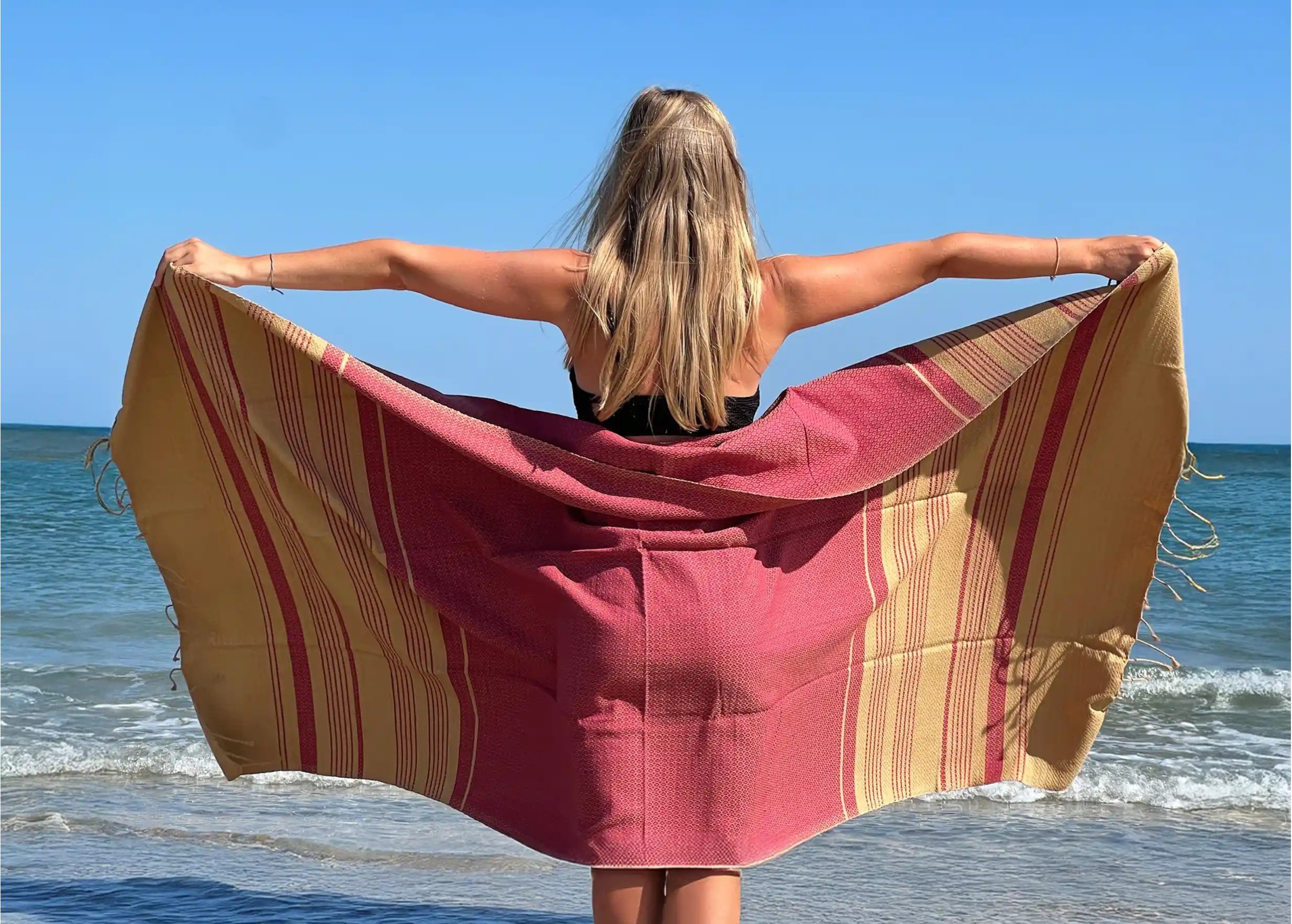 Woman on the beach seen from behind, wearing a red ochre fouta made from organic cotton from the Sand collection