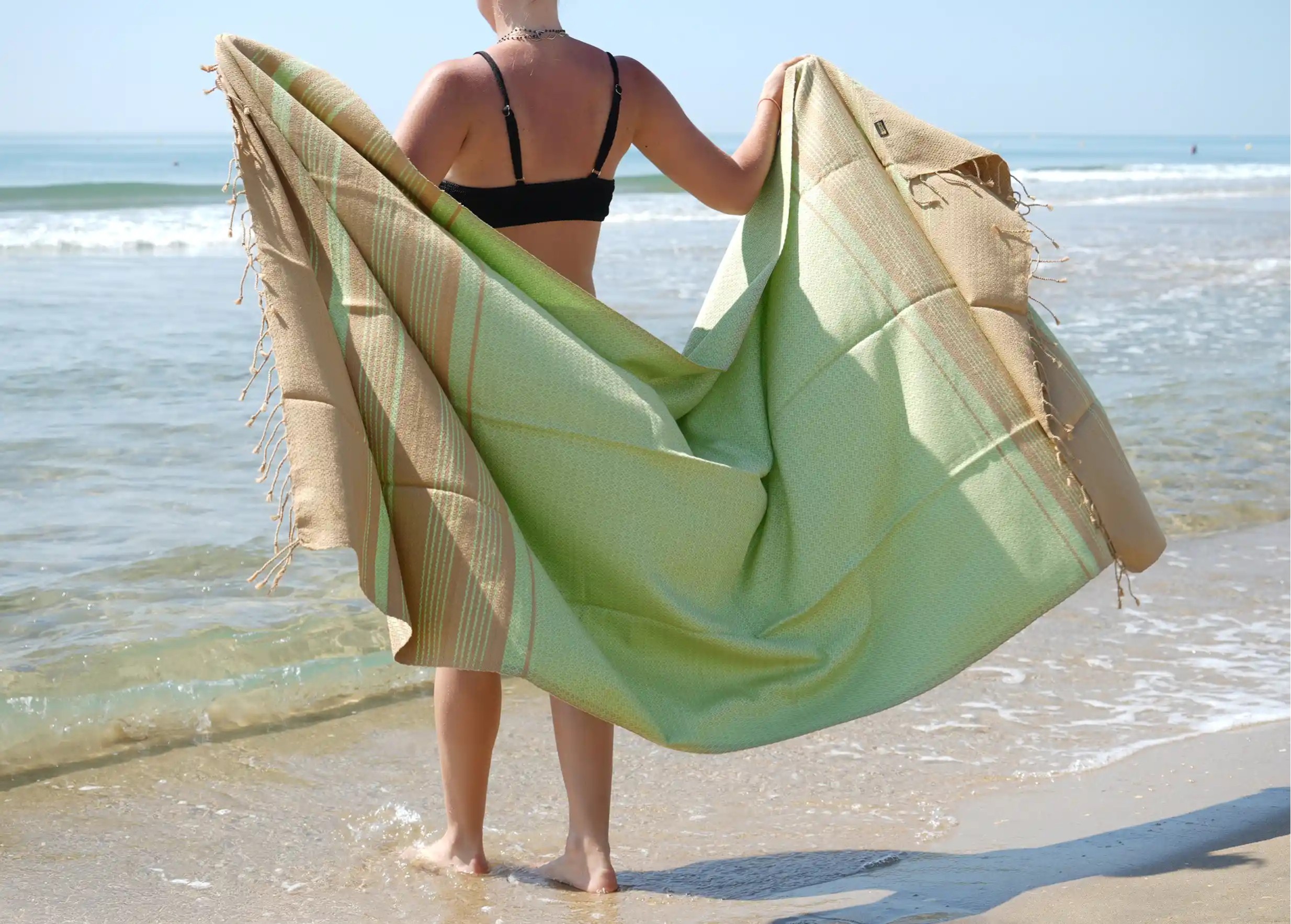 Woman seen from behind, facing the beach, holding a Karawan brand fouta with her fingertips.