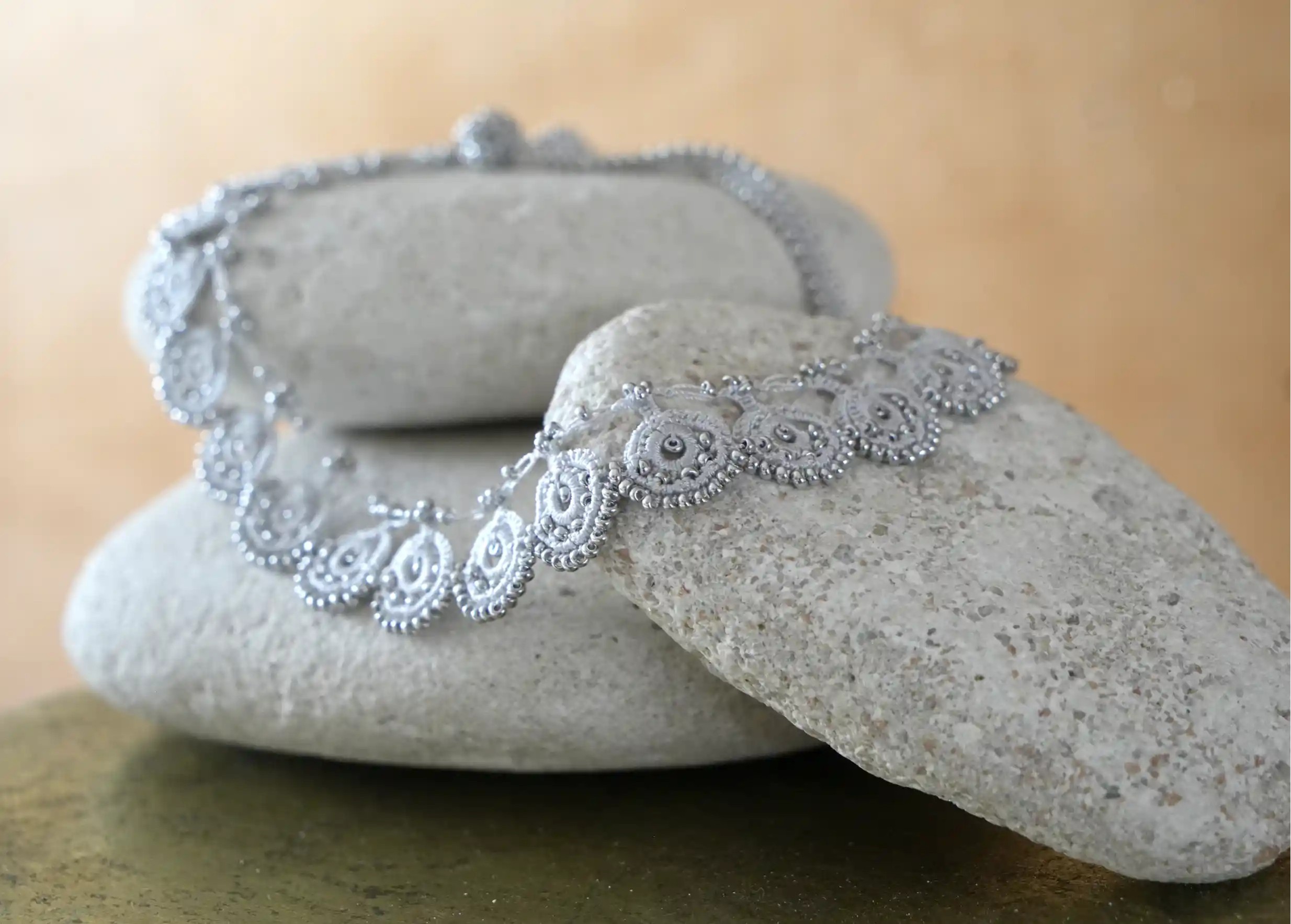 Gros plan sur le motif en dentelle du collier Eda coloris gris argent posé sur des galets gris clair