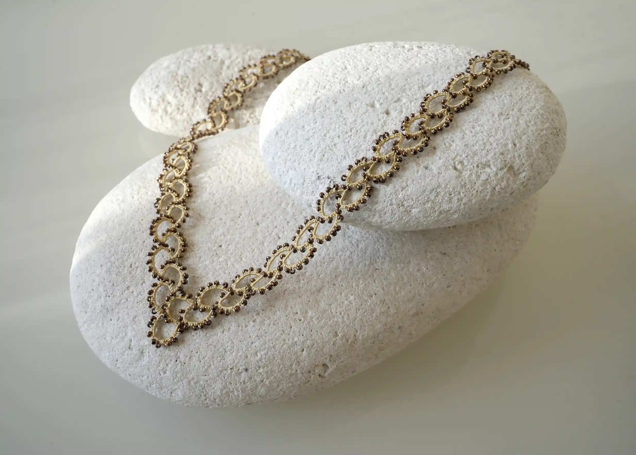 Collier en dentelle et perles réalisé à la main, modèle Seed et coloris beige et or, posé sur un galet