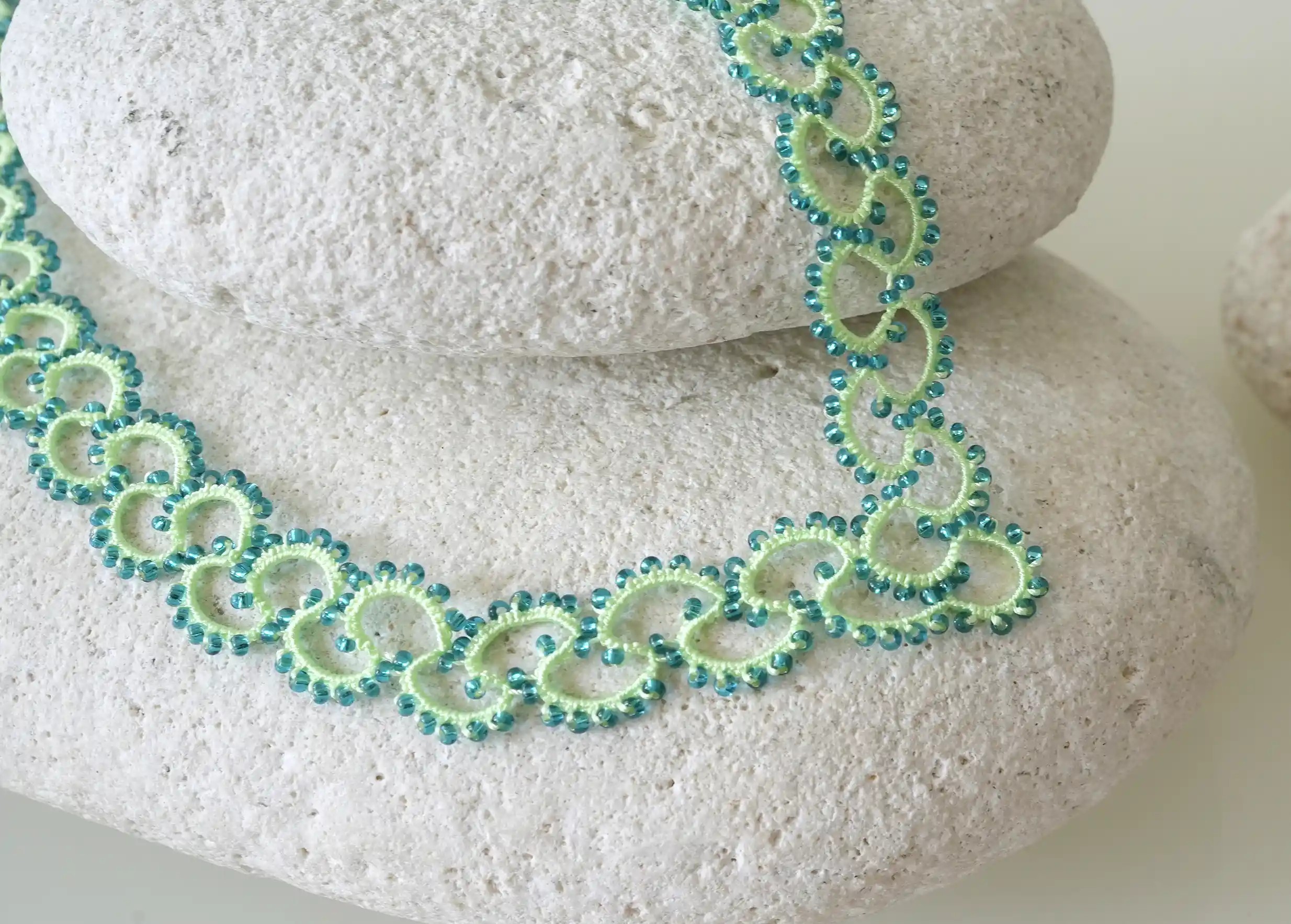 Collier en dentelle et perles réalisé à la main, modèle Seed et coloris vert émeraude, posé sur un galet