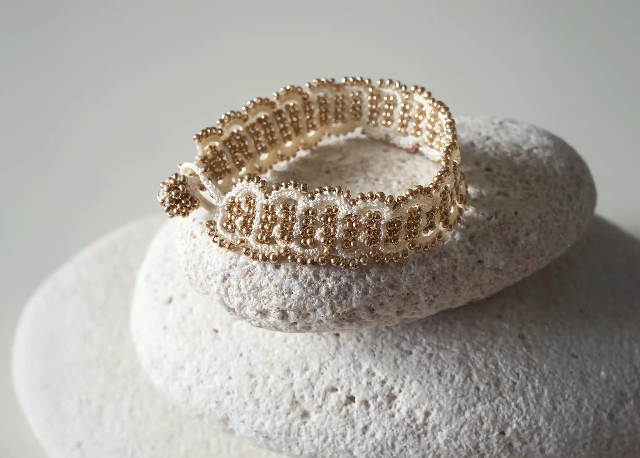 Bracelet en fil et perles coloris ivoire et or posé sur un galet, modèle Bark