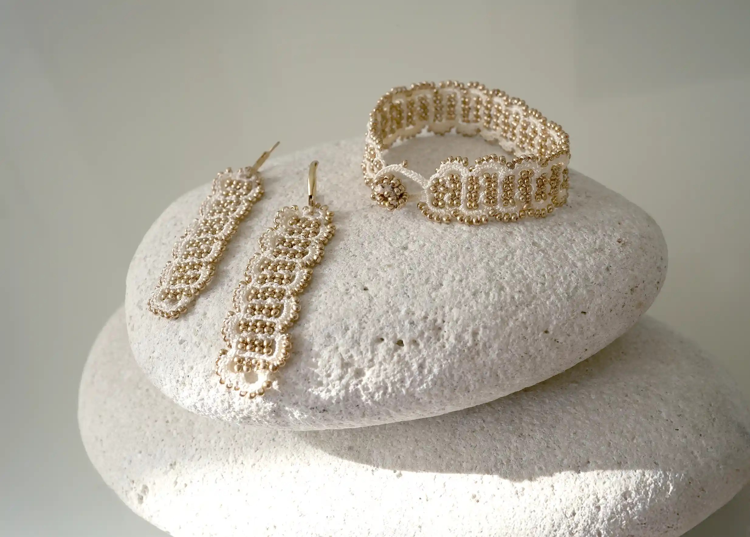 Ensemble de boucle et bracelet en fil et perles coloris ivoire et or posés sur un galet, modèle Bark