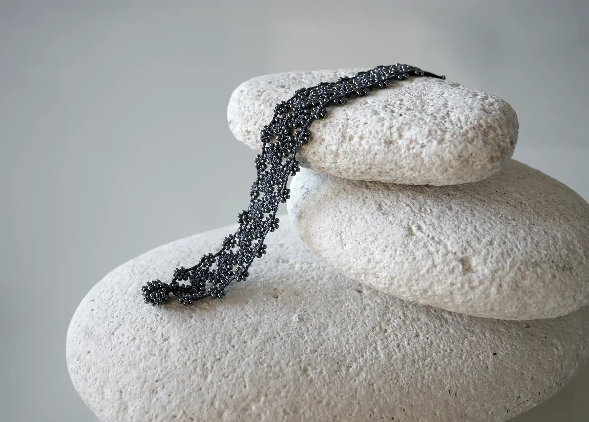 Bracelet textile en fil et perles, avec des motifs graphiques, modèle Ribbon coloris bleu ardoise, posé sur un galet