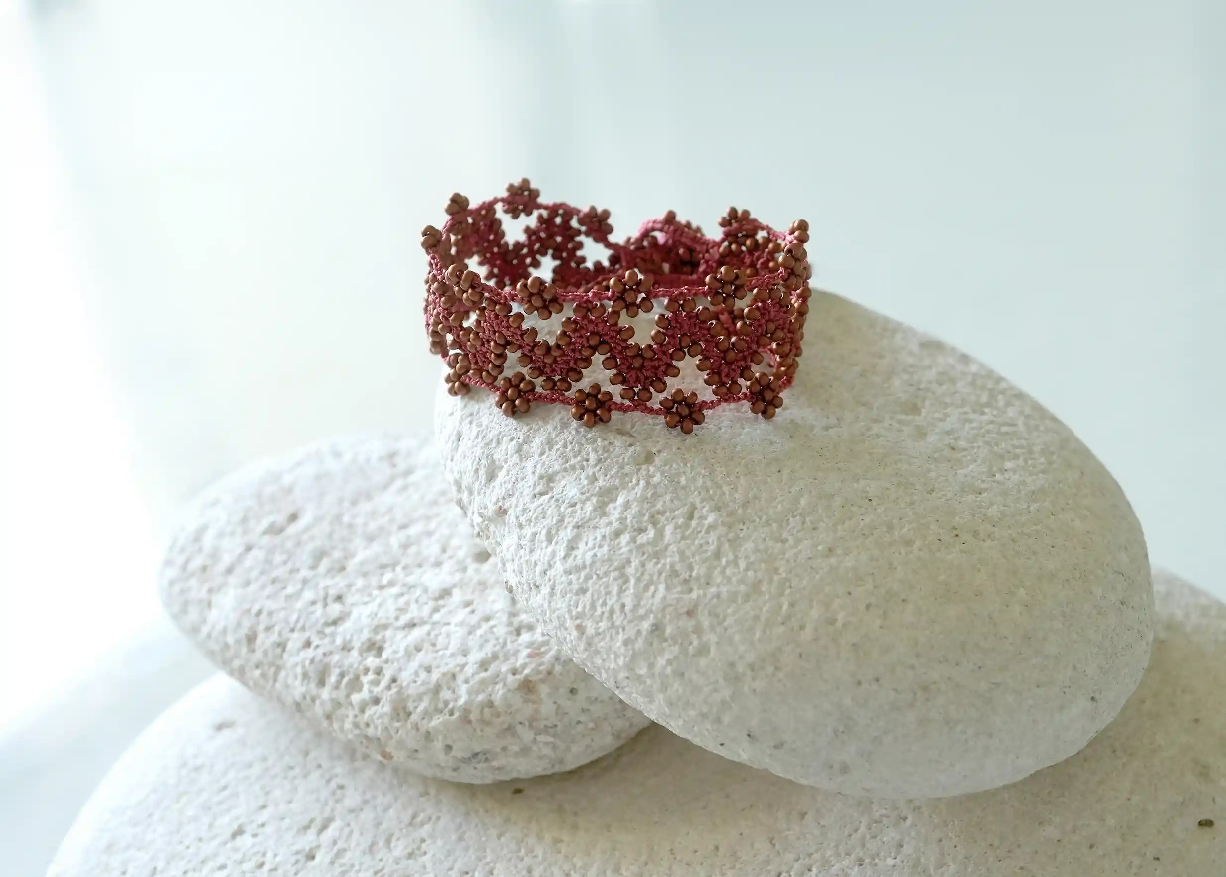 Textile bracelet made of thread and beads, with graphic patterns, Ribbon model in matt copper colour, placed on a pebble