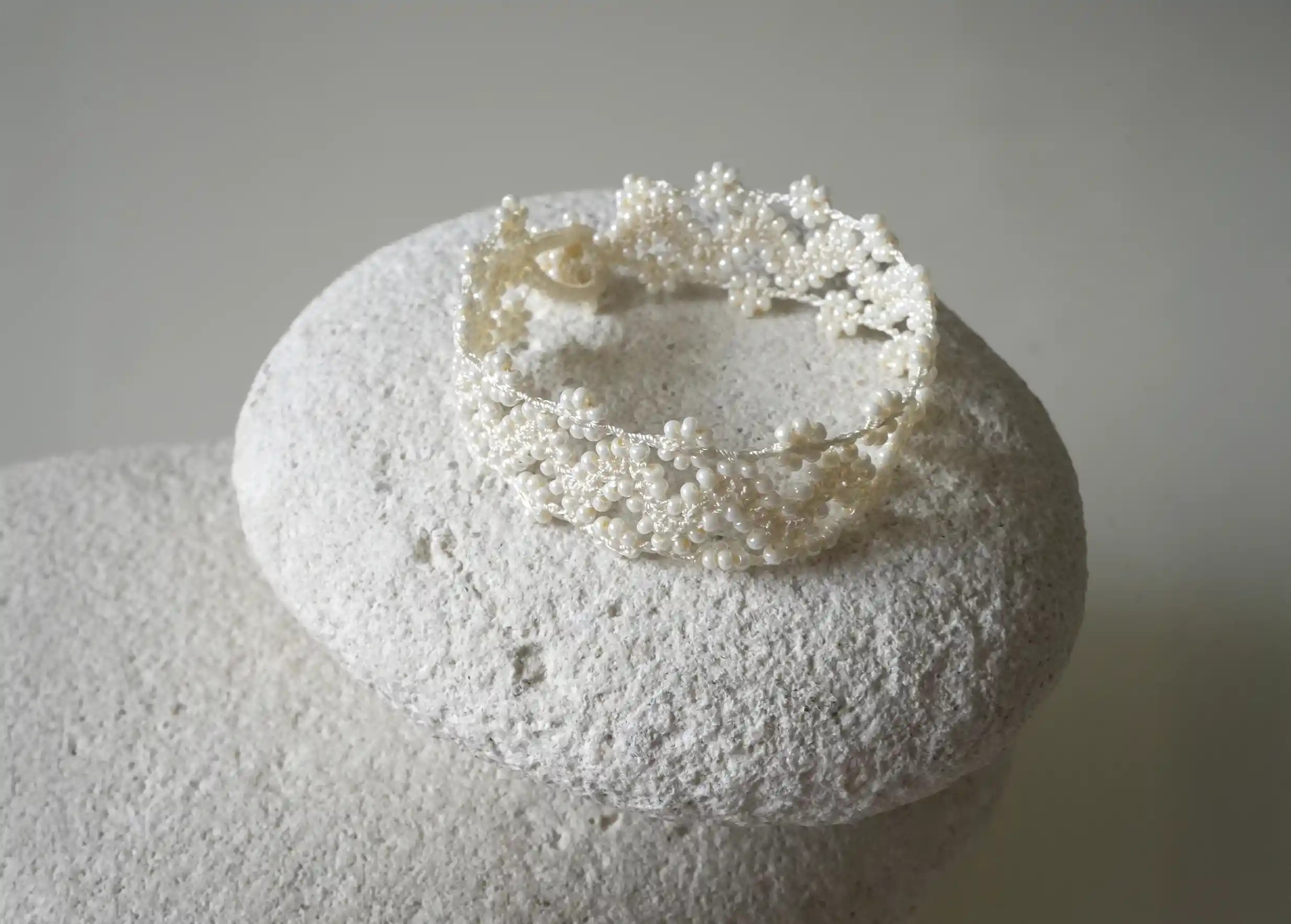 Textile bracelet made of thread and beads, with graphic patterns, Ribbon model in light ivory colour, placed on a pebble
