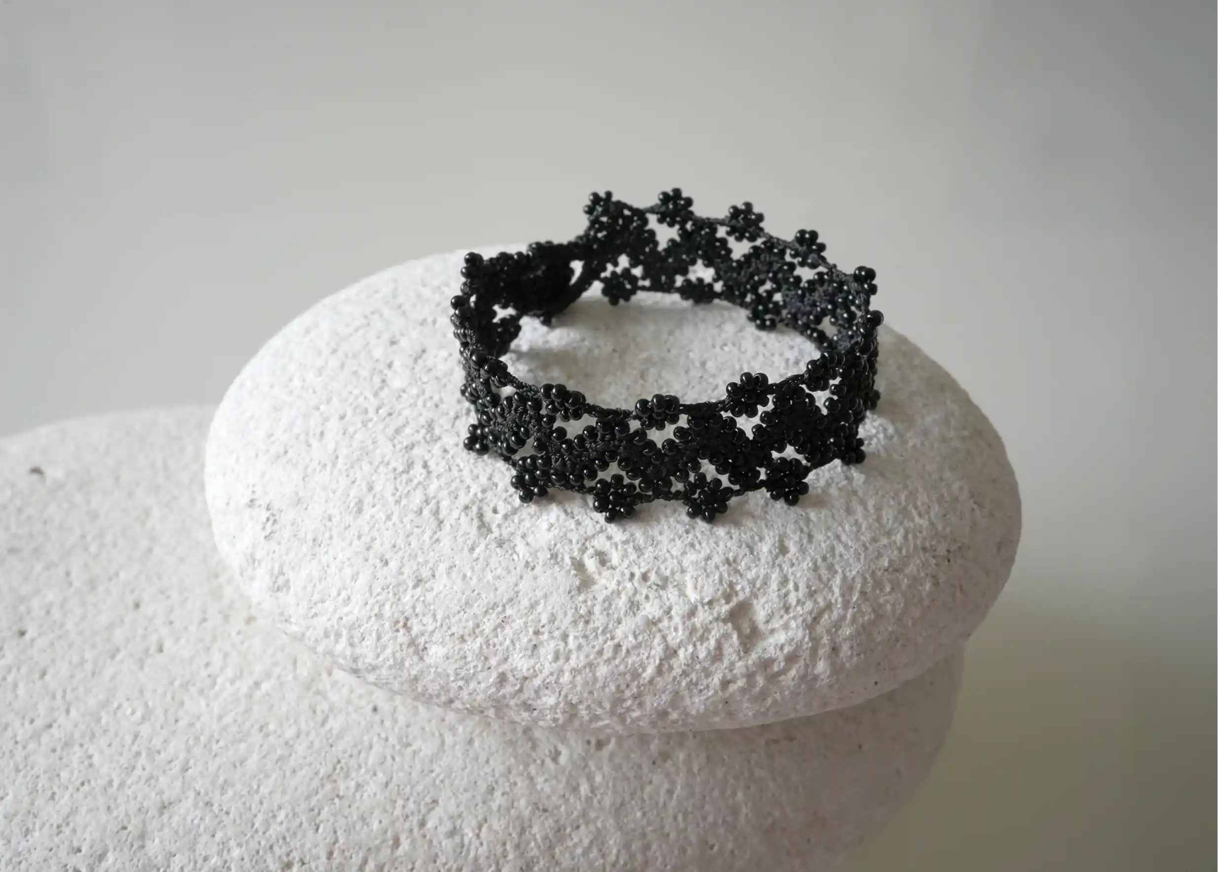 Bracelet textile en fil et perles, avec des motifs graphiques, modèle Ribbon coloris noir, posé sur un galet