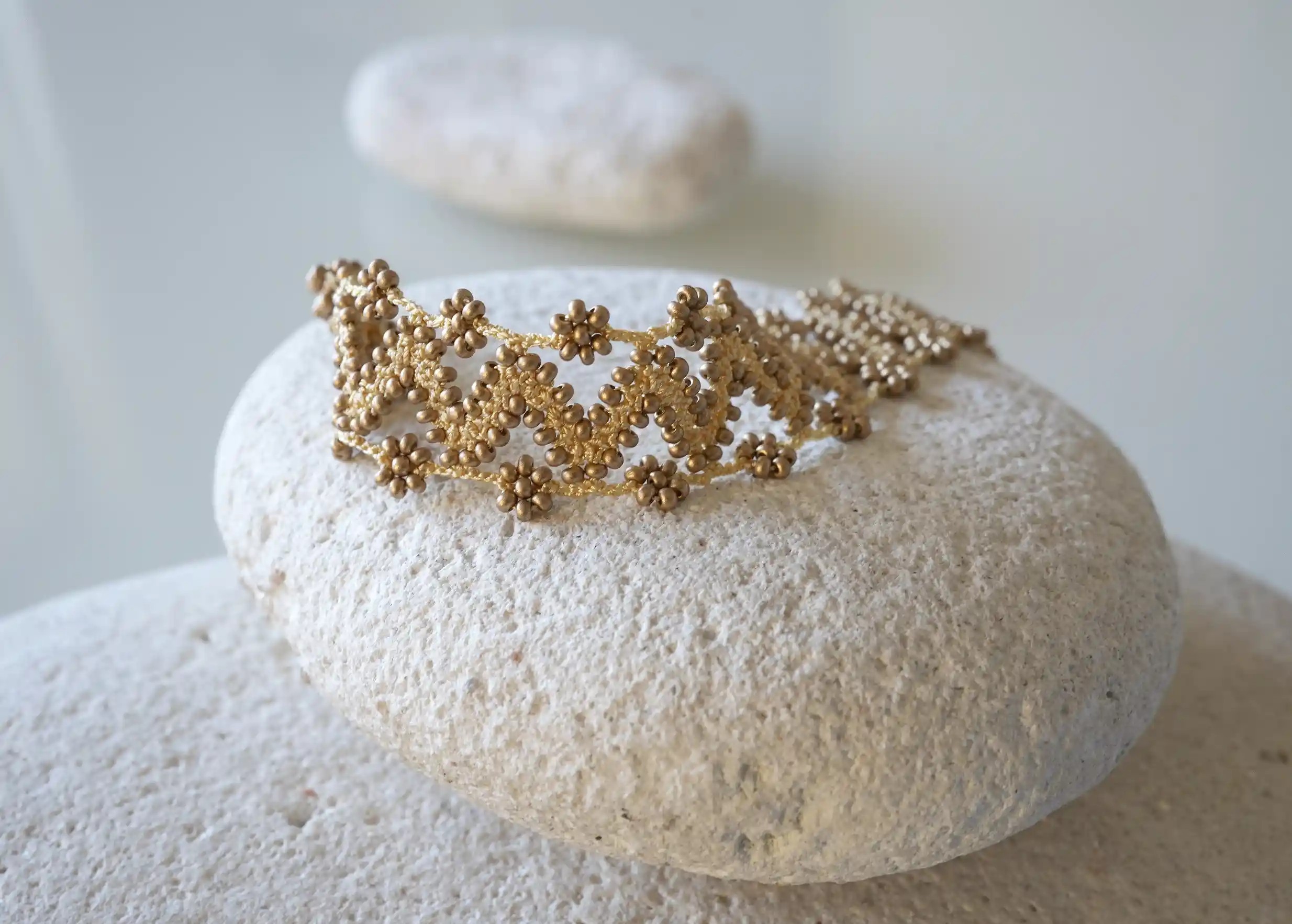 Textile bracelet made of thread and beads, with graphic patterns, Ribbon model in matt gold, placed on a pebble