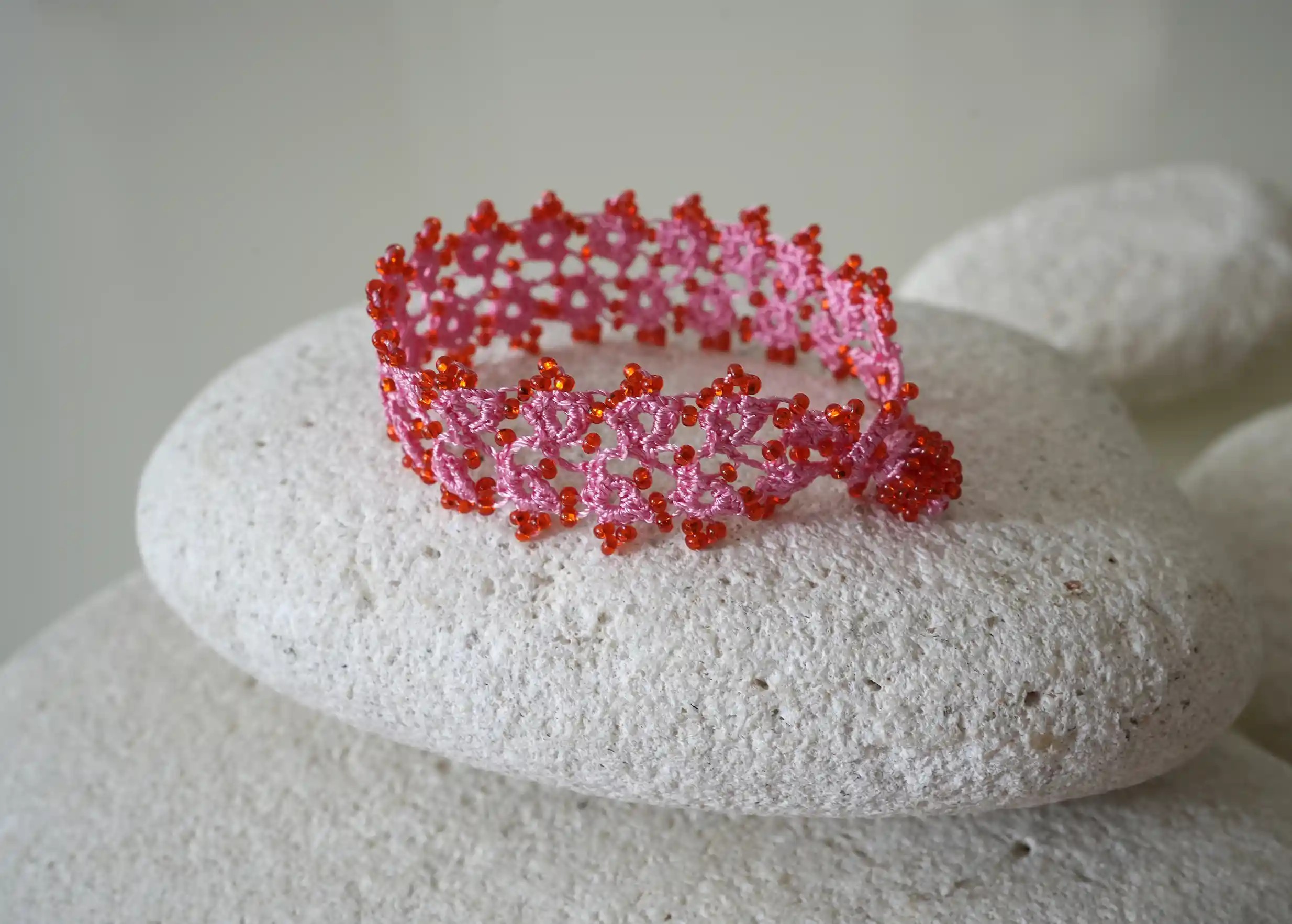 Handmade lace textile bracelet, Husnu, OYA collection, pink and red colours placed on a pebble