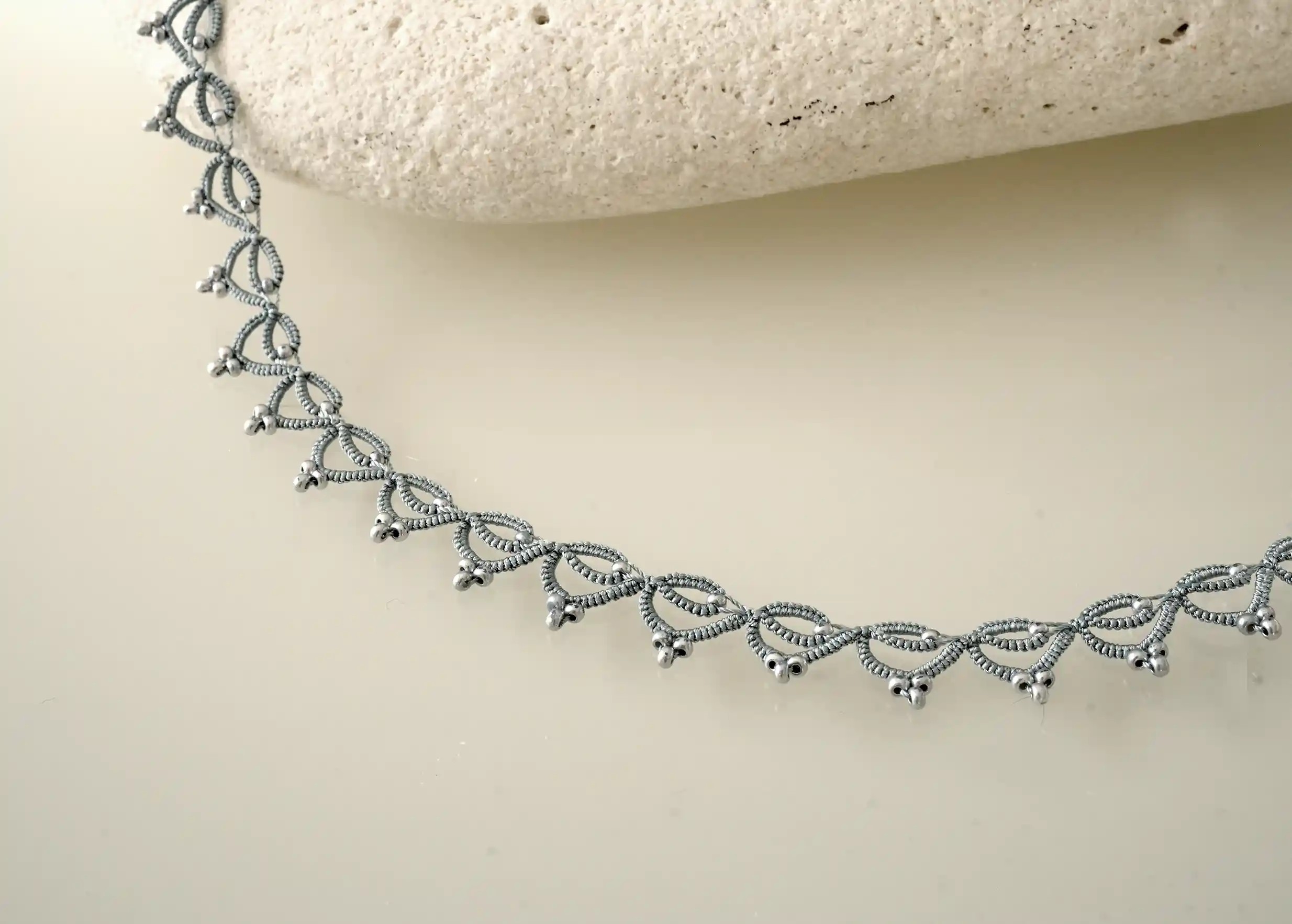 Handmade choker necklace made from thread and beads near a pebble, Lotus Chain in glacier blue and silver